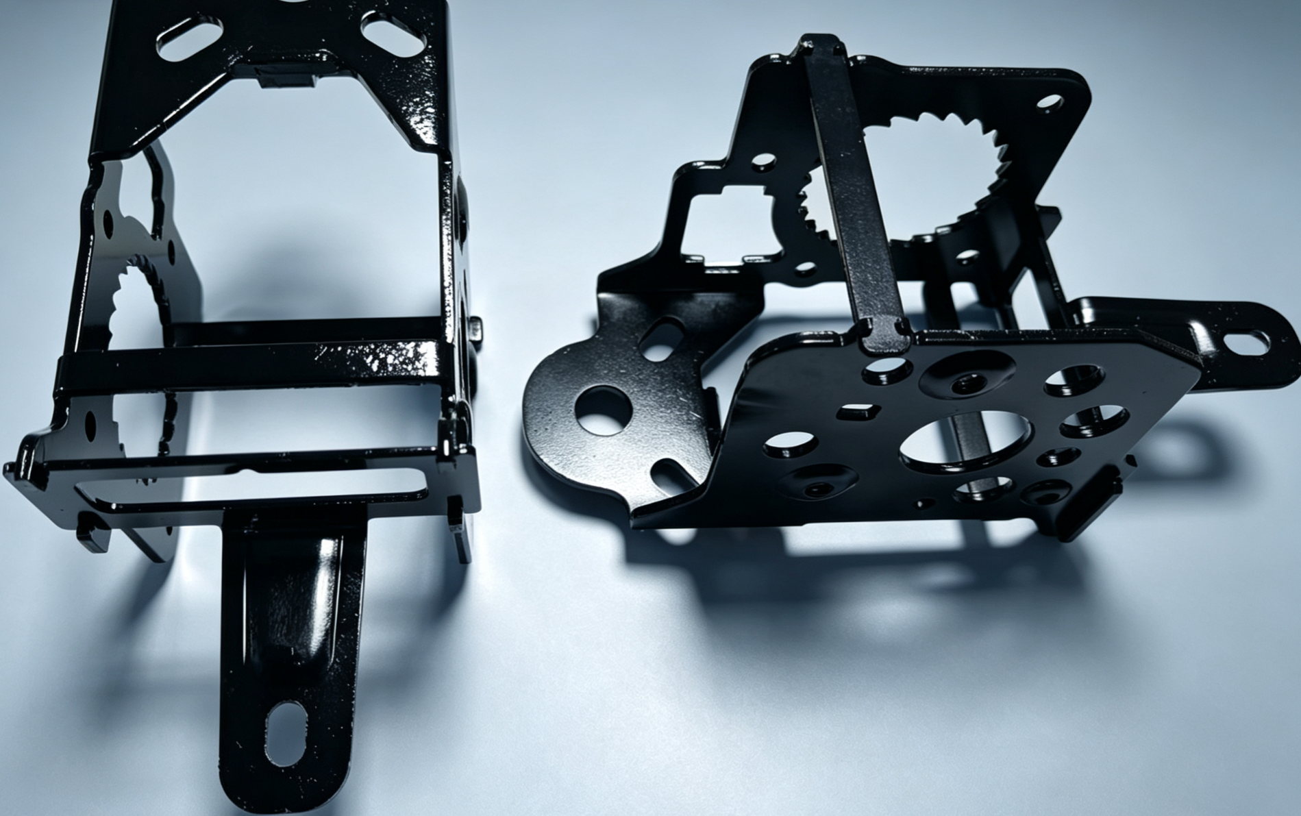 Two black coated automotive seat frame brackets with serrated cut-outs, slots and mounting holes